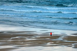 Lone figure in red on the quiet seashore watching the waves.